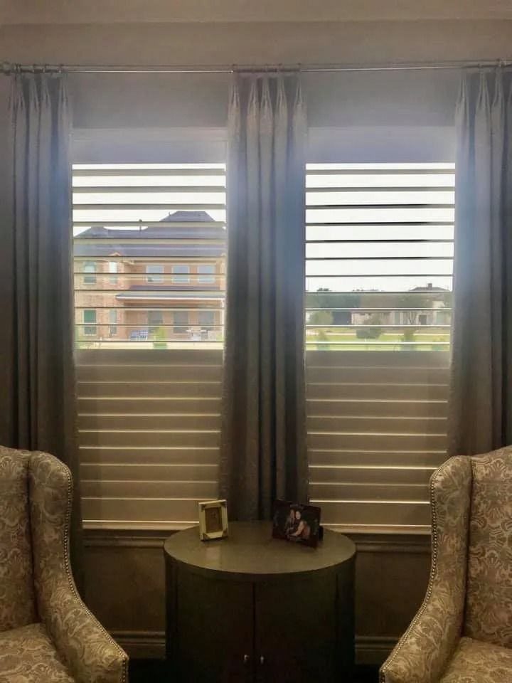 Two windows with blinds and curtains, flanked by patterned armchairs, a side table, and buildings in the background.