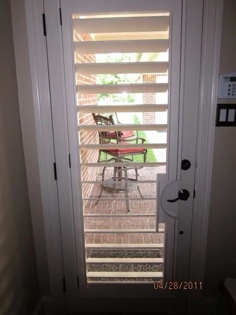 White door with shutters revealing a patio with red chairs and a table.