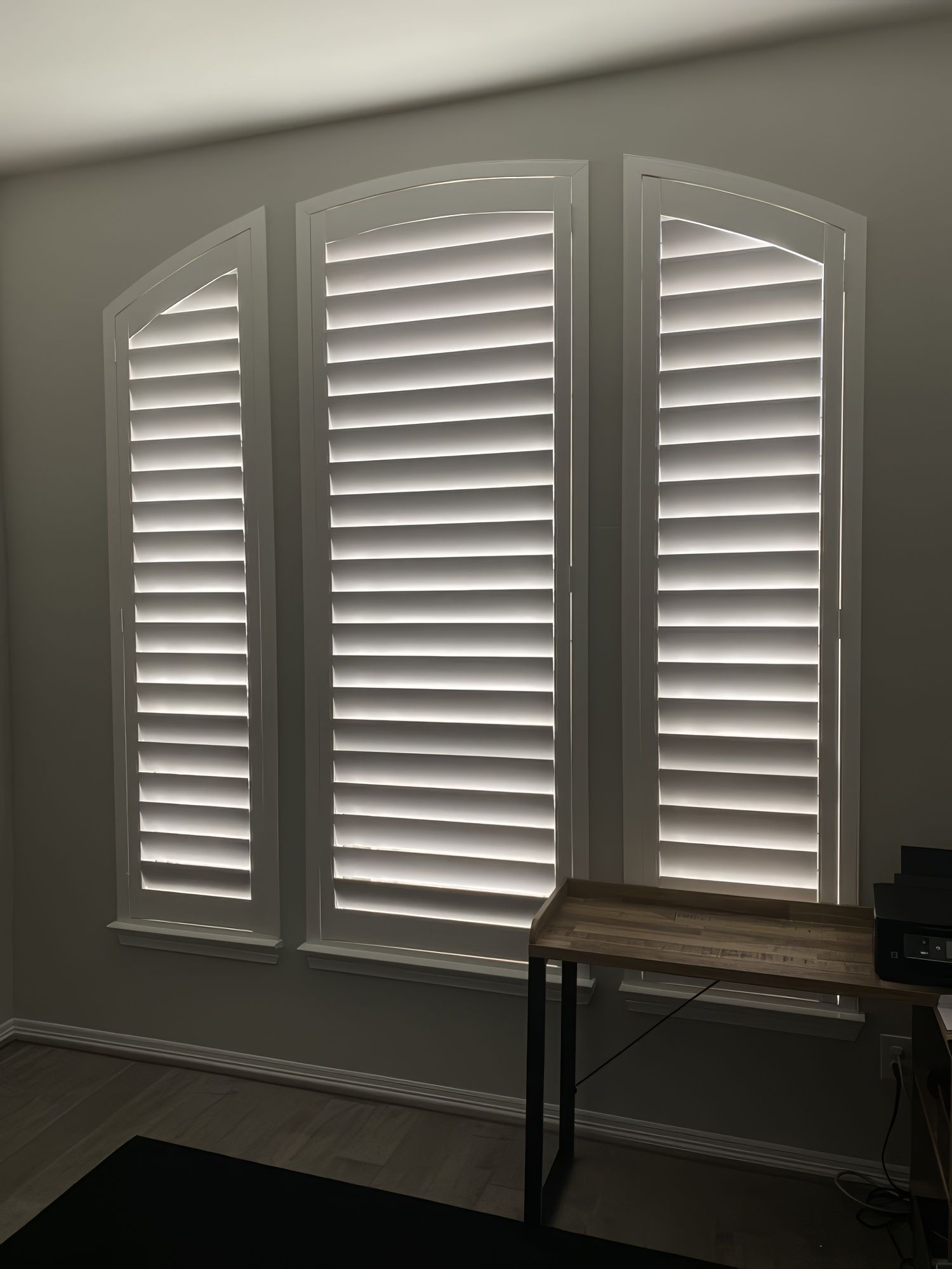 White shuttered window with an arched top, partially open, letting in light. A small desk sits below.