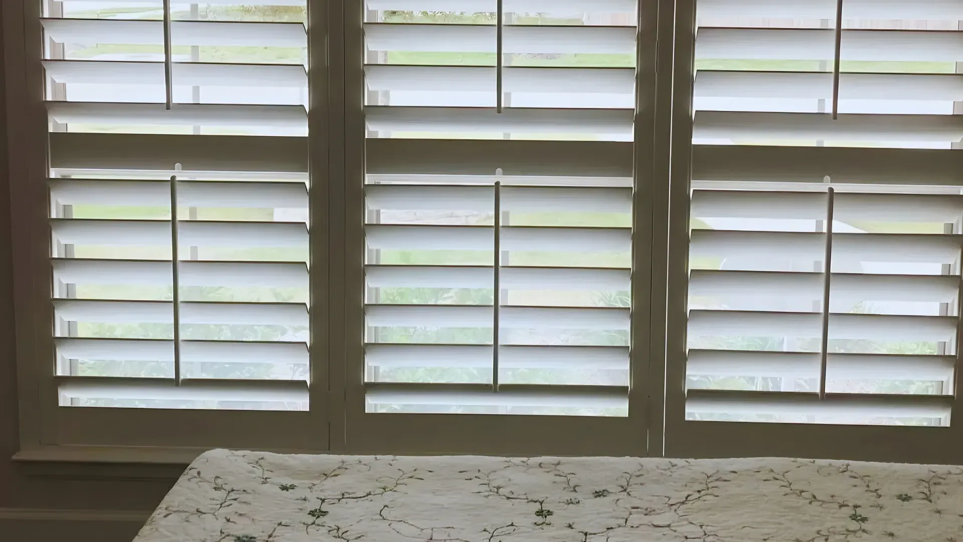 Closed white window shutters, natural light filtering through, atop a lace-covered table.