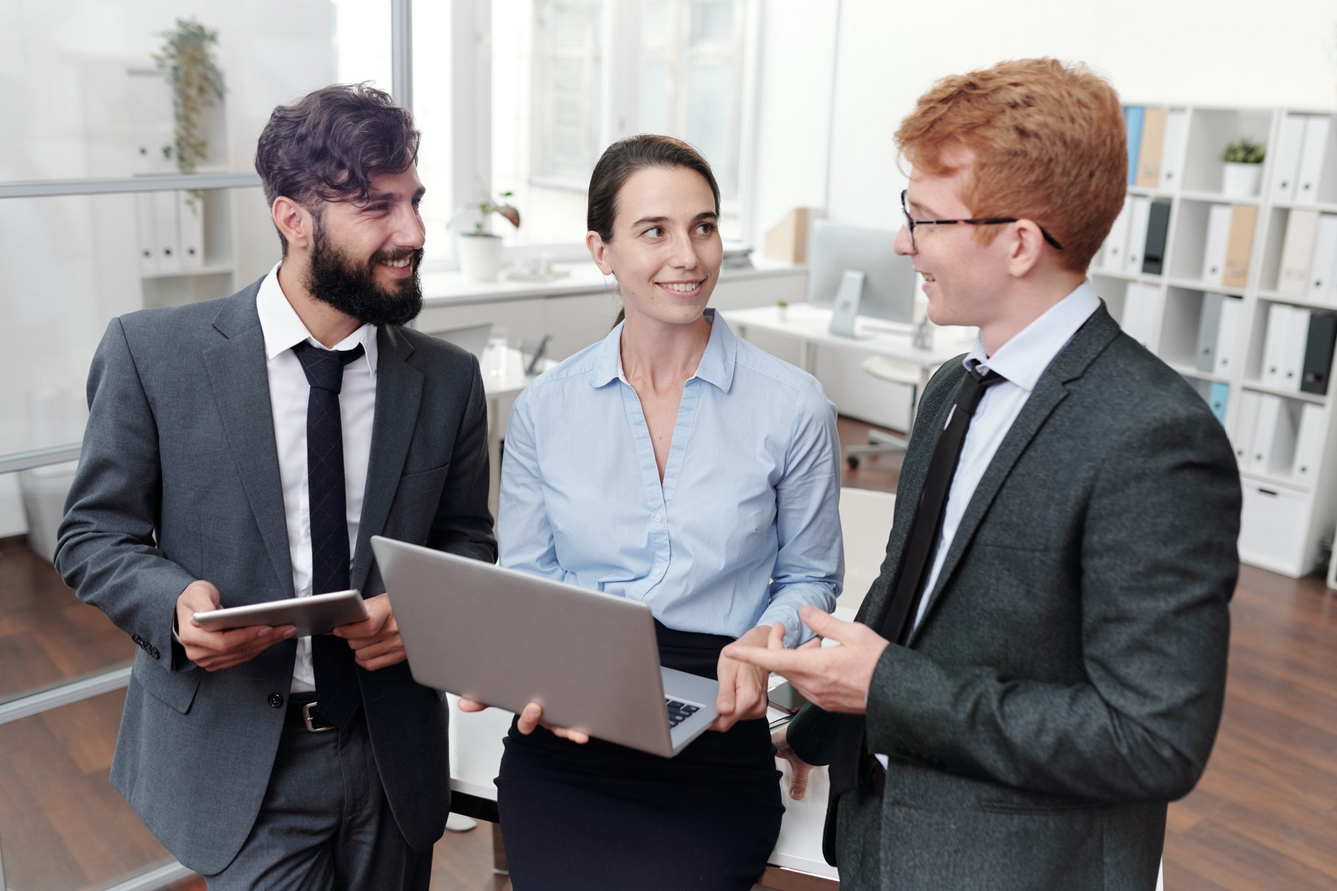 Three people in suits discussing, two holding a laptop and tablet, in an office.