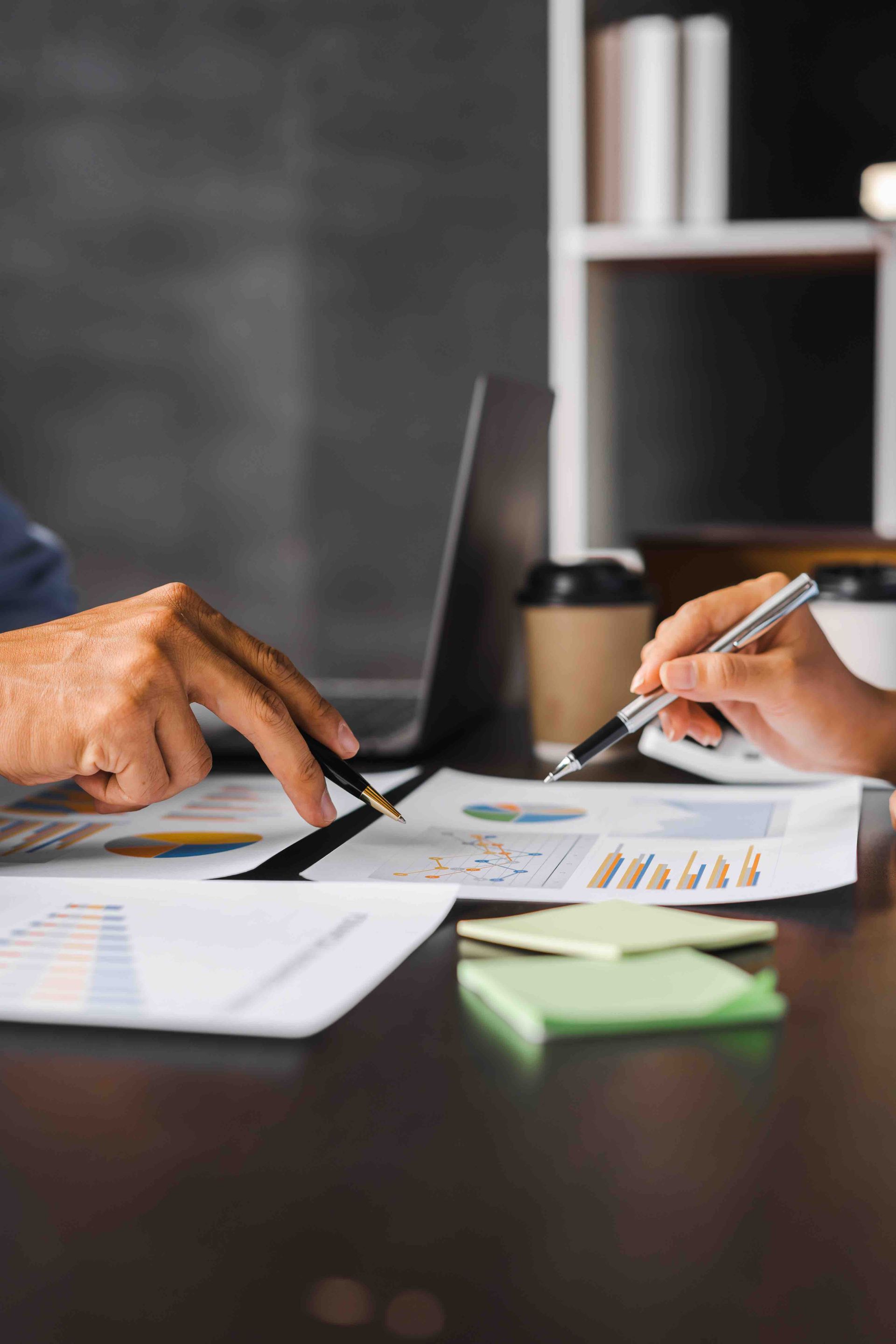 Two people pointing at charts and documents on a desk with a laptop and coffee cups.