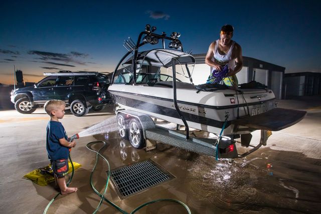 A man is washing a boat with a hose while a little boy watches in the parking lot of a storage garage unit Tamworth.
