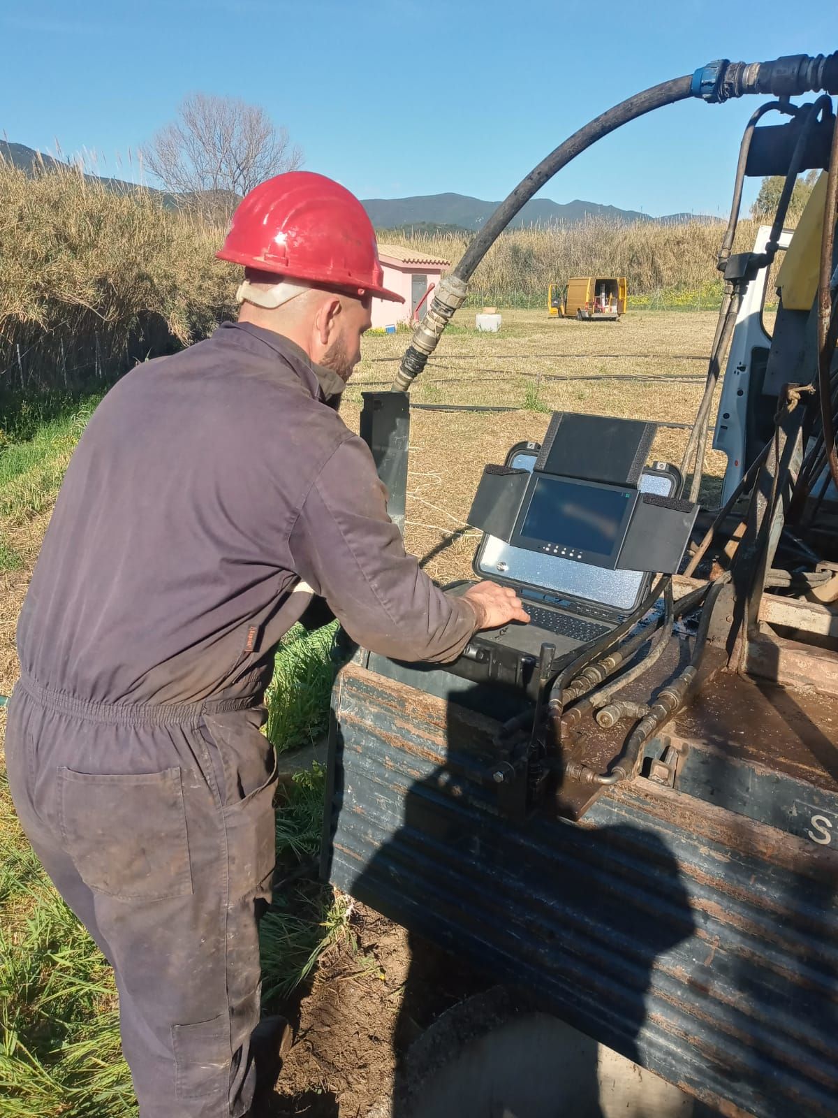 Un uomo con un casco rosso usa un computer portatile vicino a un'attrezzatura di perforazione all'aperto, probabilmente in un cantiere edile.