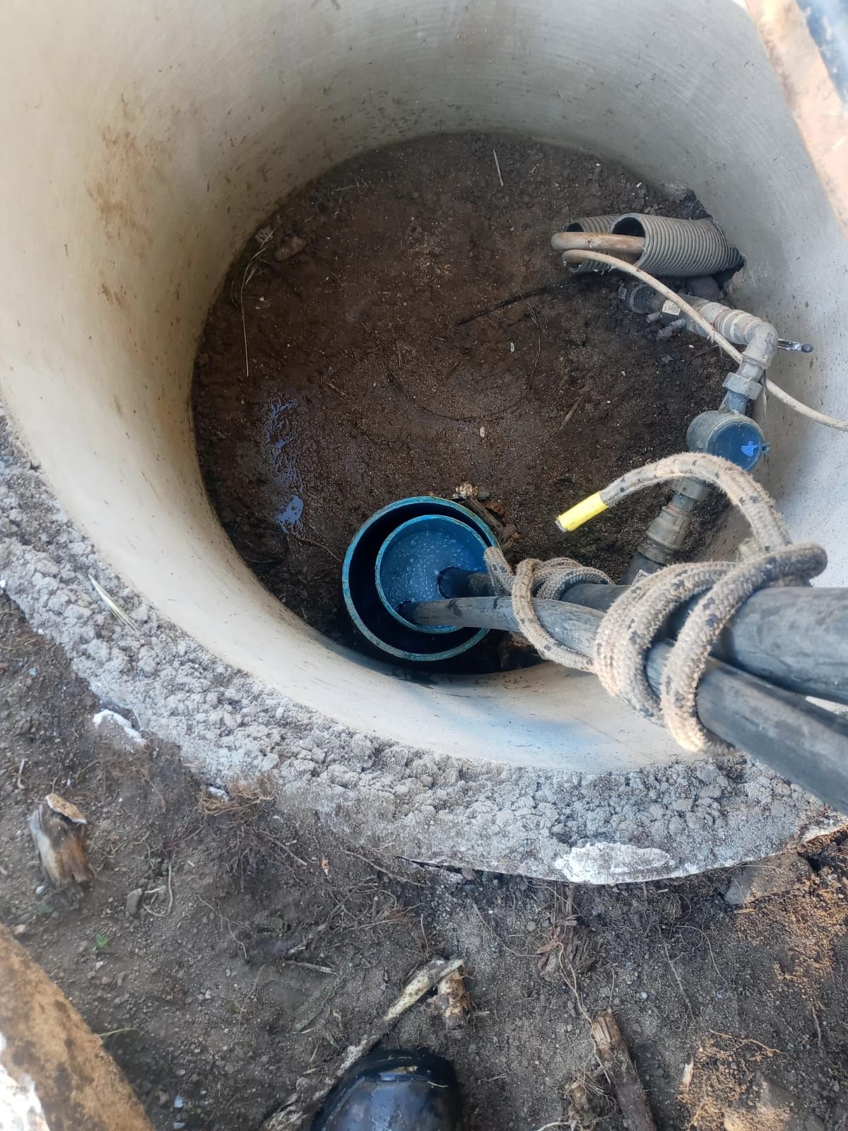 Vista dall'alto di un pozzo con una pompa e cavi di alimentazione all'interno. L'interno del pozzo è sporco e detriti.