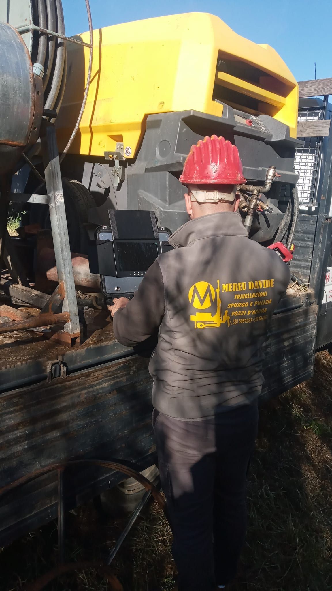 Operaio con casco rosso e abiti da lavoro vicino a un'attrezzatura di perforazione; macchina gialla, ambiente esterno.