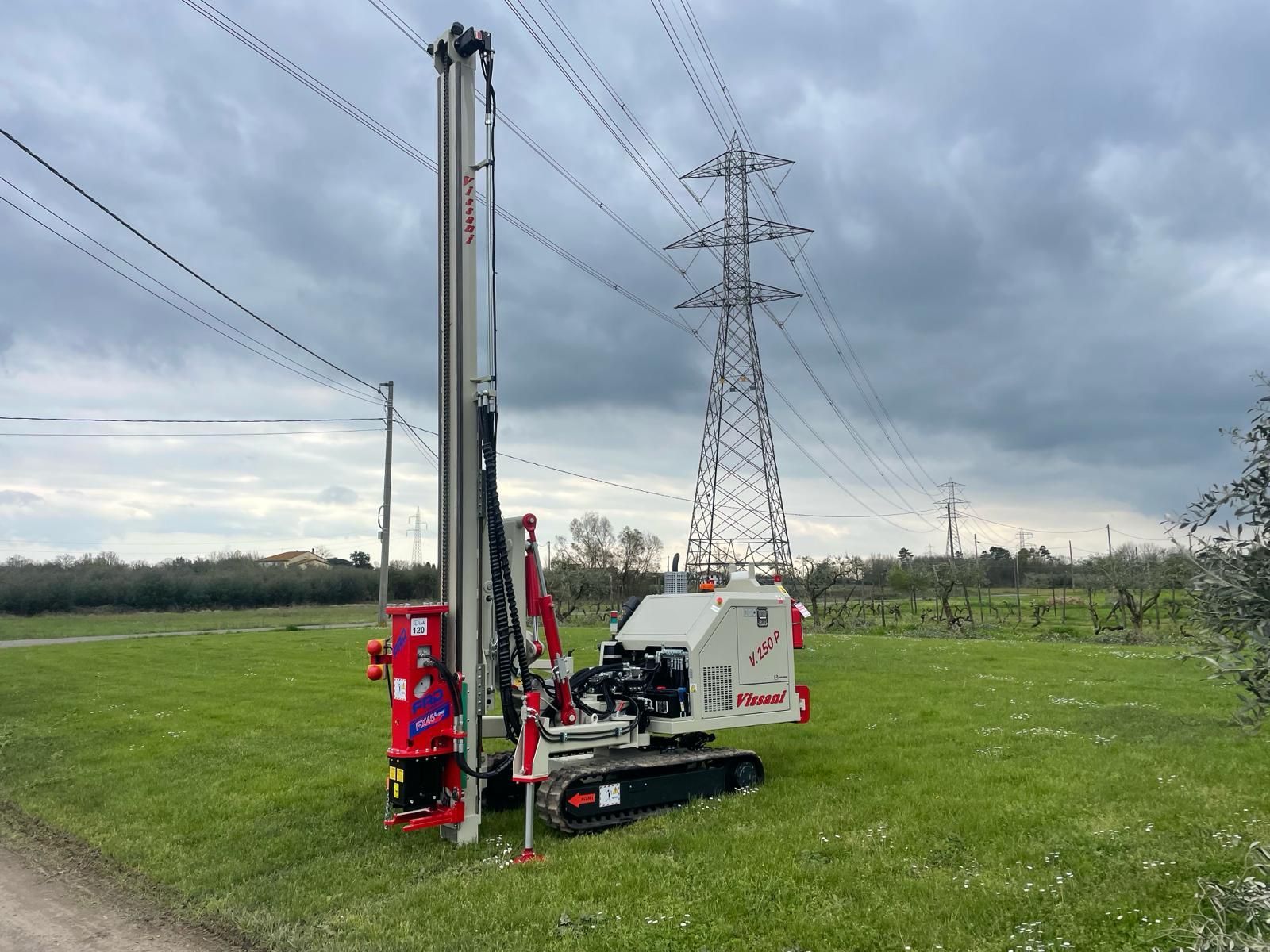 Perforatrice su cingoli in un campo, vicino a linee elettriche e a una torre elettrica, sotto un cielo nuvoloso.