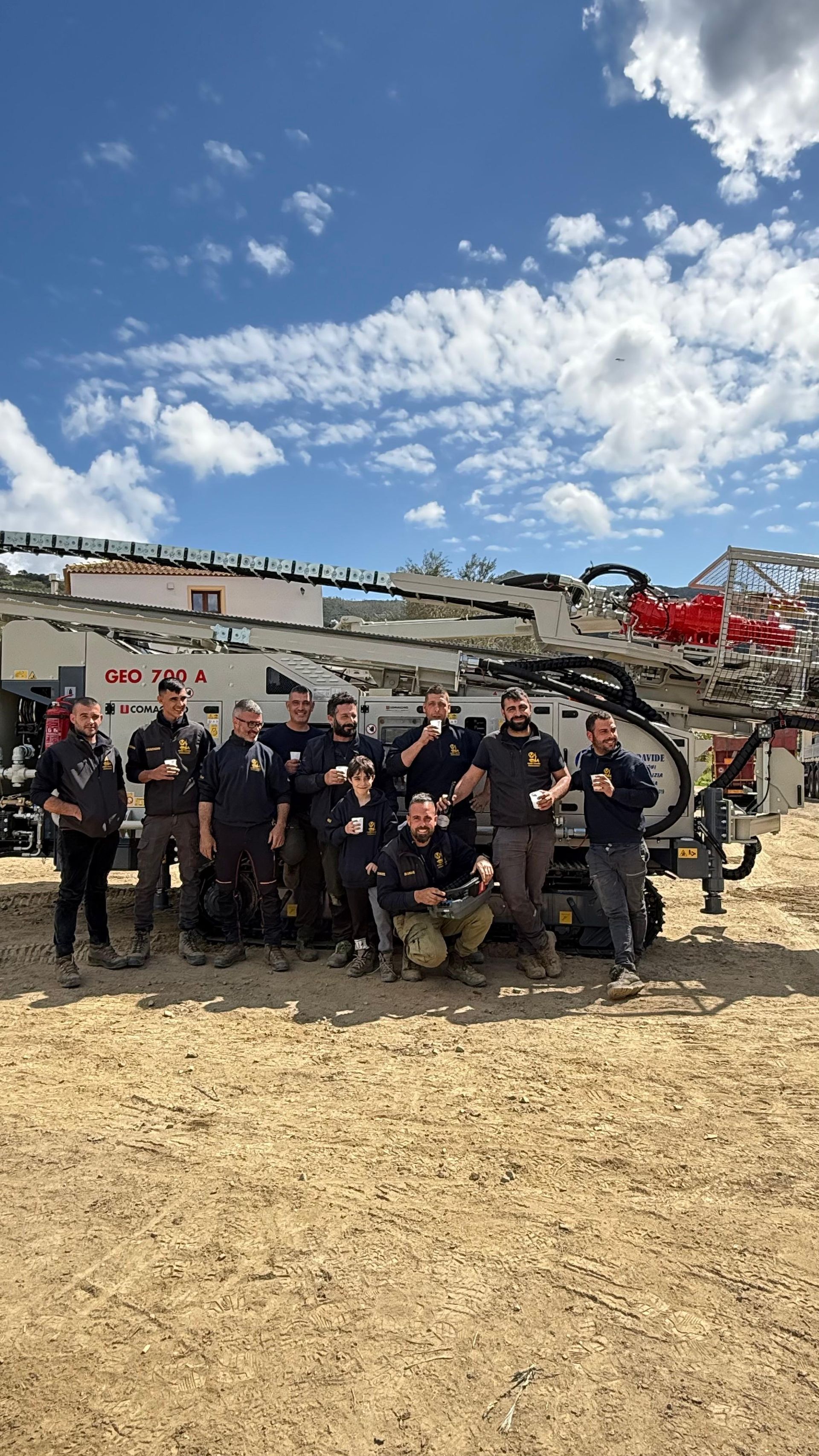 Gruppo di persone in posa con grandi macchinari industriali bianchi all'aperto sotto un cielo azzurro.