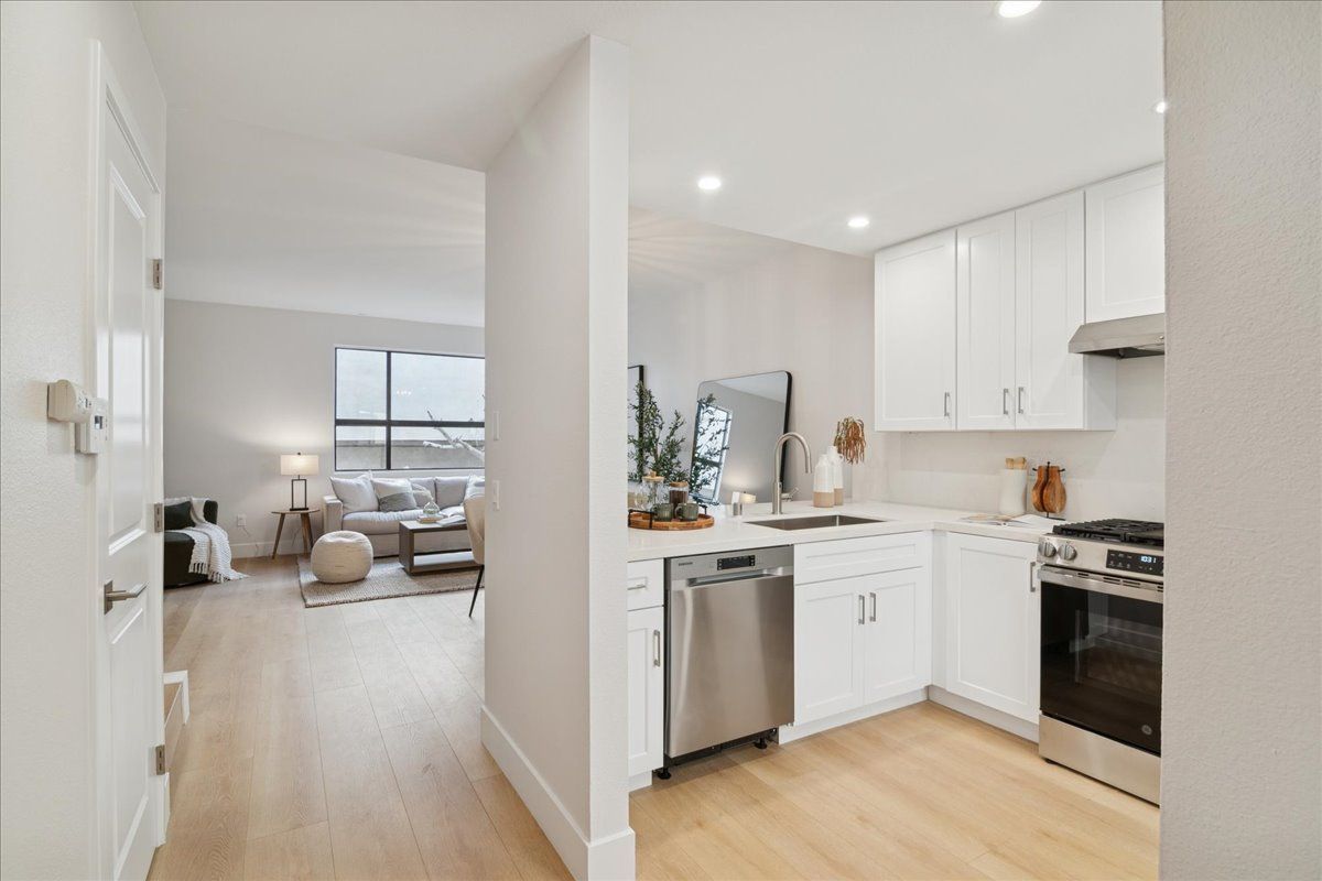 Bright open kitchen with white cabinets beside a light-filled living room and hallway