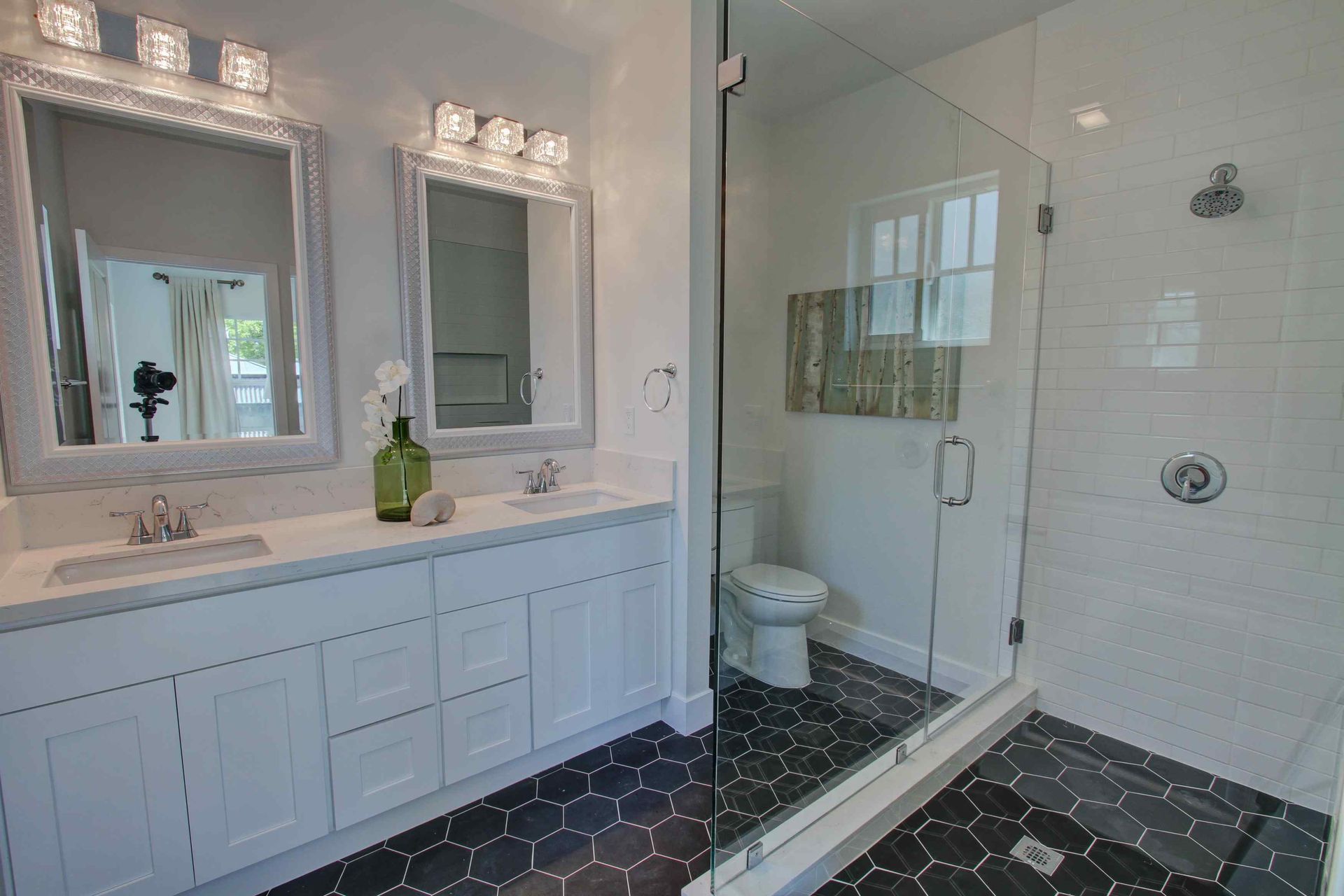 Bright bathroom with double vanity, large mirror, glass shower, and dark patterned floor tiles.