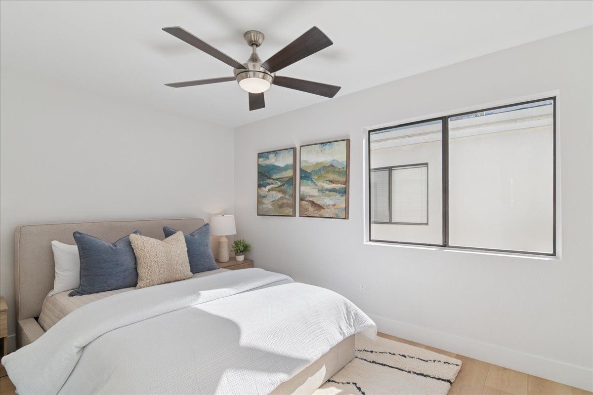 Bright bedroom with a beige bed, blue pillows, ceiling fan, wall art, and two windows