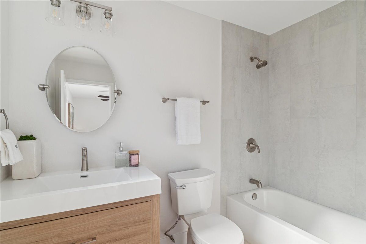 Bright modern bathroom with white tub, toilet, vanity, round mirror, and light wood cabinet.