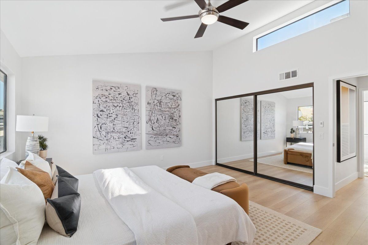 Bright modern bedroom with white bedding, mirrored closet, ceiling fan, and abstract wall art
