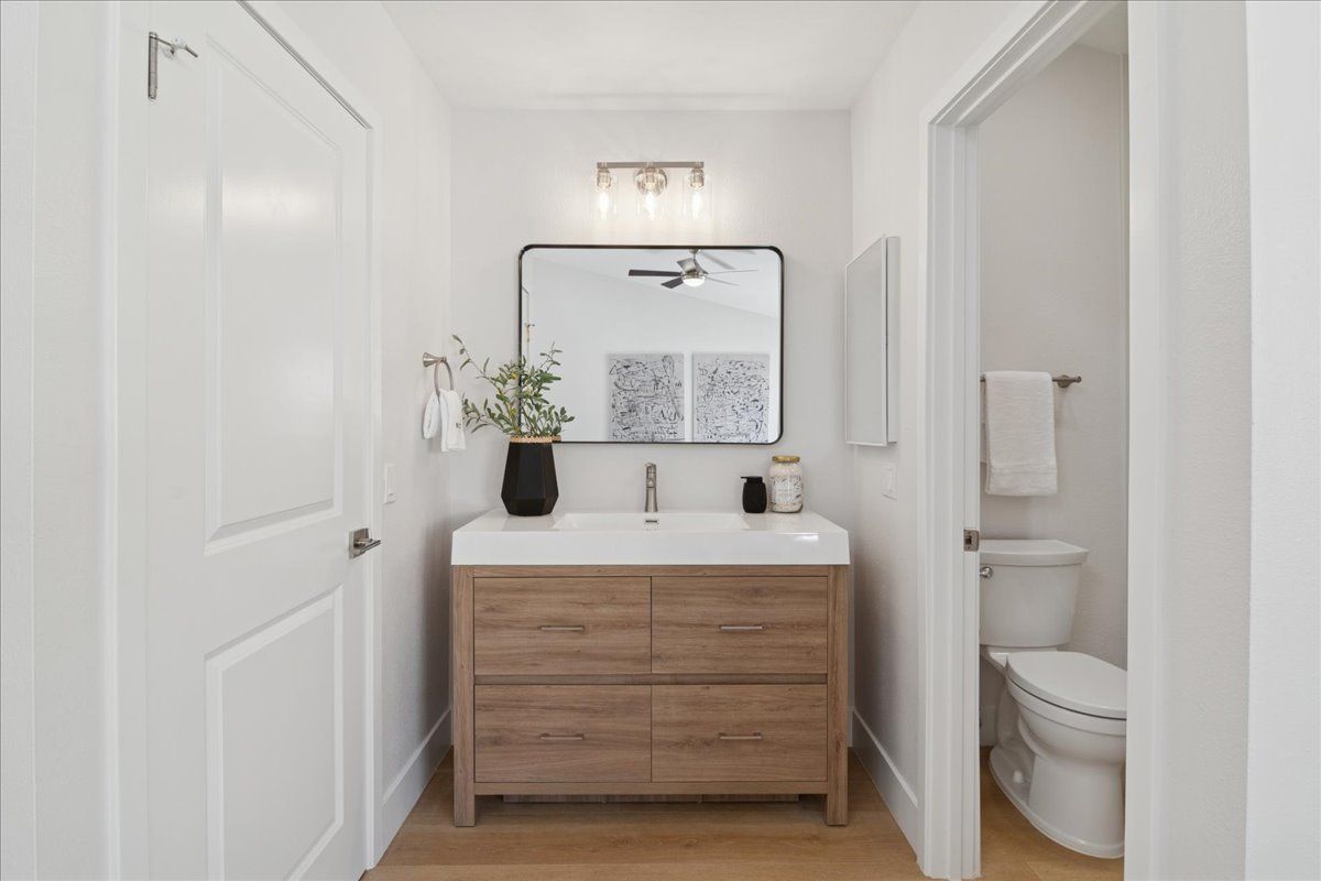 Bright bathroom with white walls, wood vanity, mirror, and a toilet in a small adjoining room.