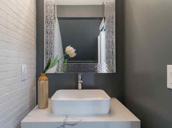A modern bathroom with a white sink, flower, and decorative mirror.  Gray and white walls.