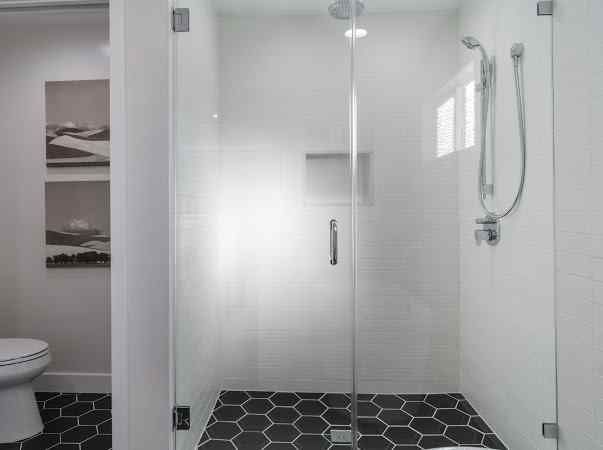Bathroom with glass shower, hexagonal black tile floor, white walls, toilet on the left, and art.