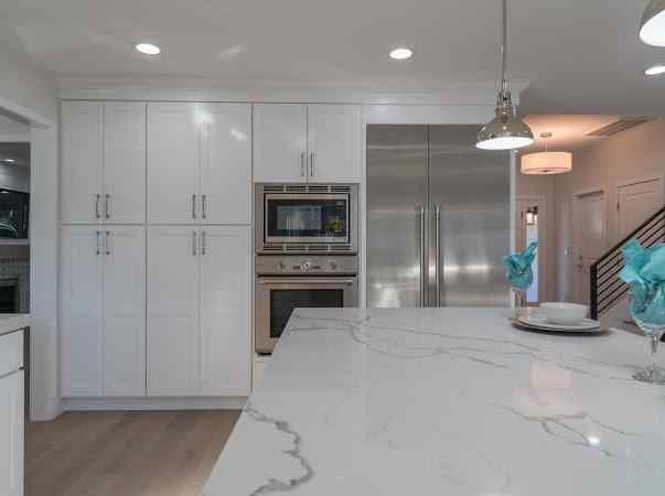 White kitchen with cabinets, built-in oven and microwave, stainless steel refrigerator, and marble countertop island.