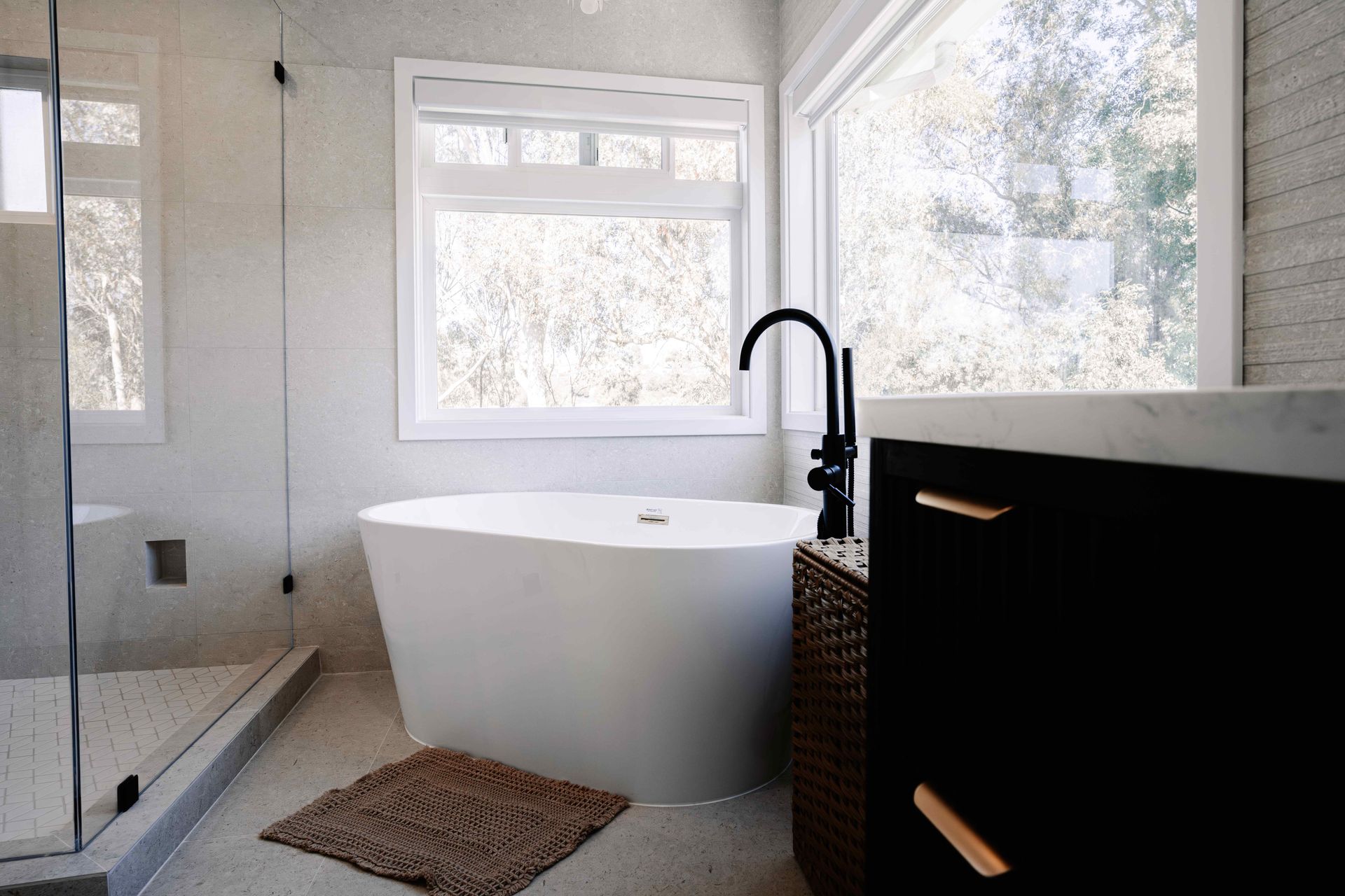 Modern bathroom with freestanding tub, large windows, black faucet, and woven rug