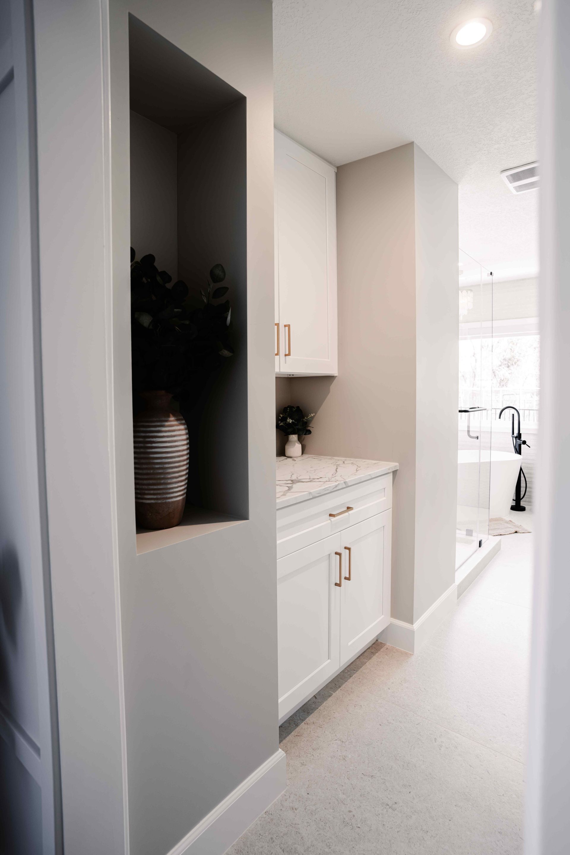 Bright narrow hallway leading to a white kitchen with cabinets, countertop, and a recessed wall niche with a plant.