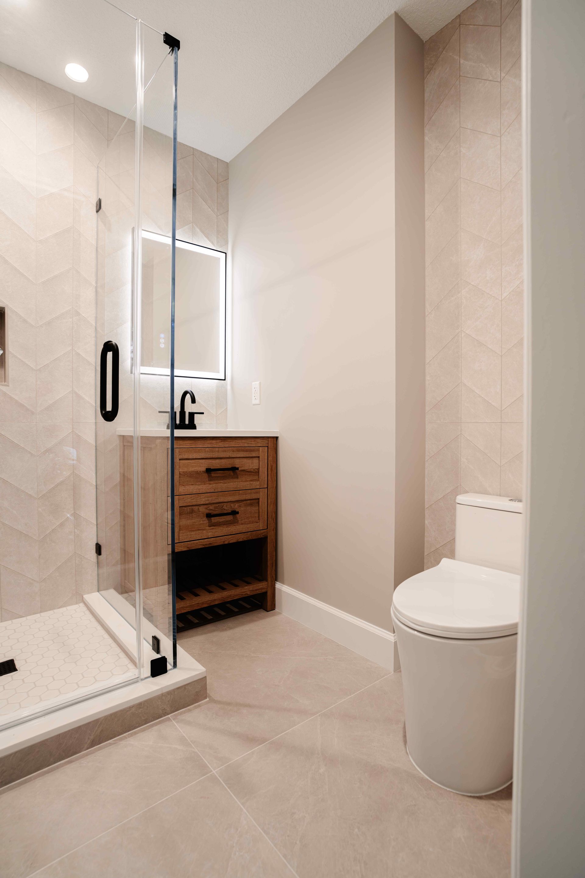 Modern beige bathroom with glass shower, wooden vanity, lit mirror, and toilet
