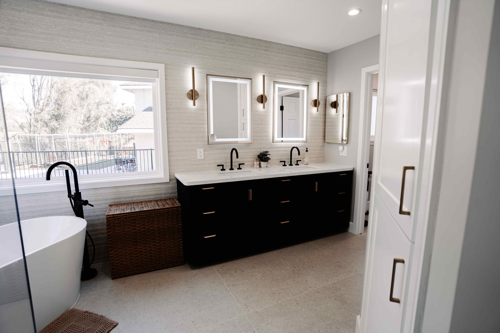 Bright bathroom with white walls, black vanity, tub, mirror cabinets, and wicker hamper by a window.