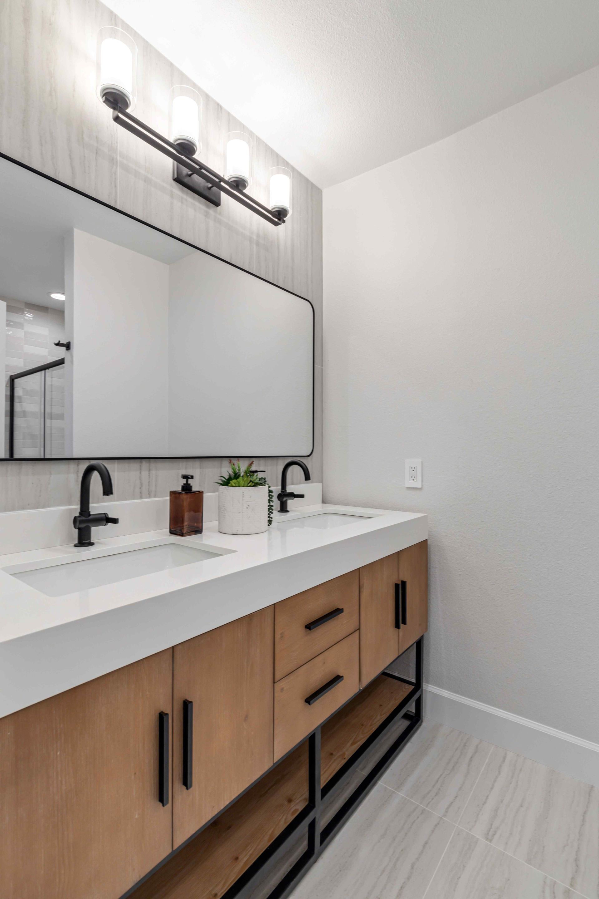 Modern bathroom vanity with wood cabinets, white countertop, black faucets, and large mirror under wall lights