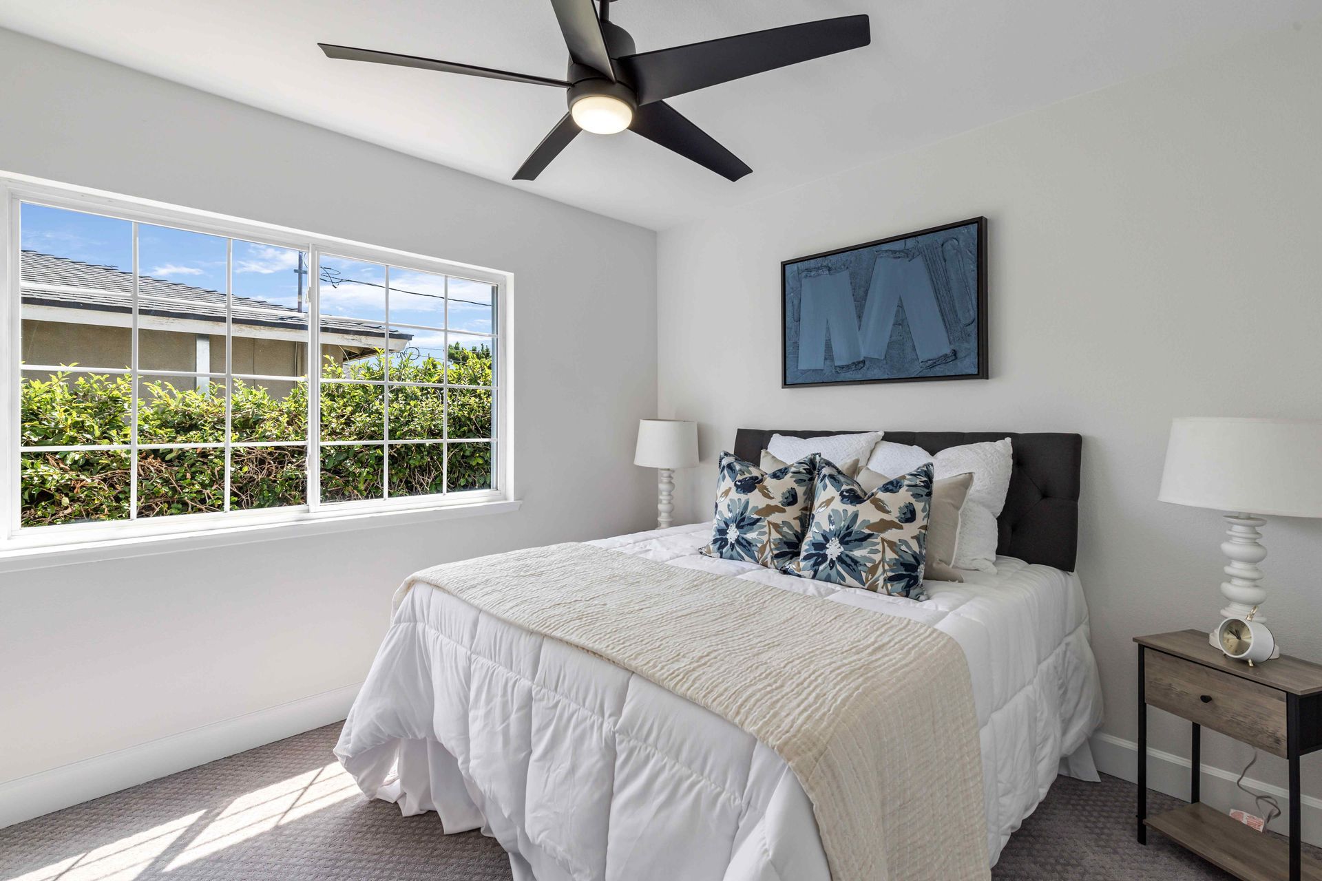 Bright bedroom with a bed, white walls, ceiling fan, and large window overlooking greenery
