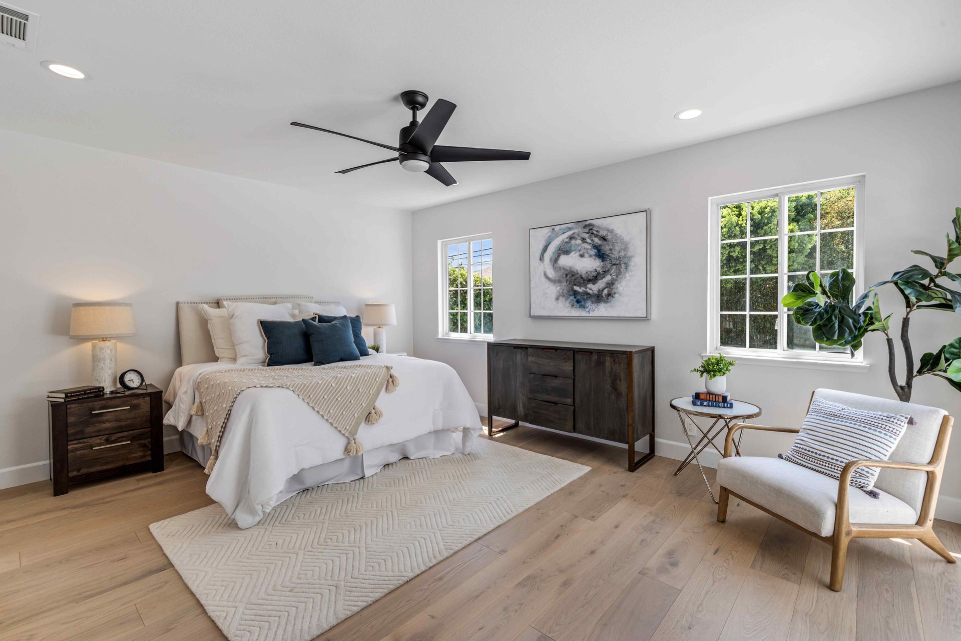 Bright bedroom with a bed, dresser, armchair, lamp, and large windows under a ceiling fan.