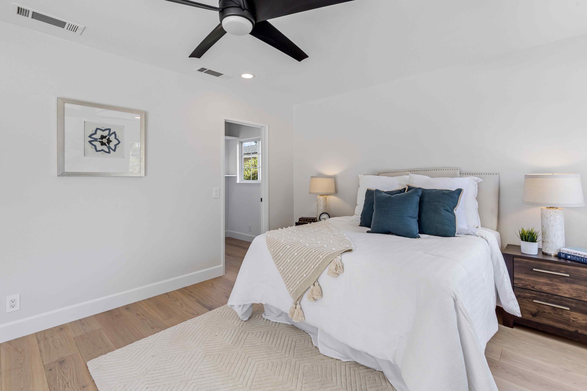 Bright bedroom with a white bed, blue pillows, nightstands, lamps, and ceiling fan in a minimalist room