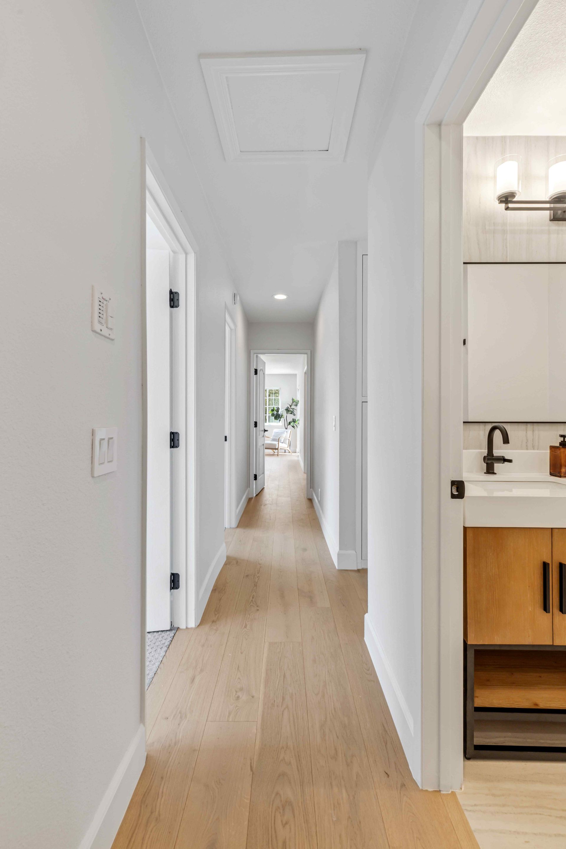 Bright white hallway with light wood floors, leading to a sunlit room and a kitchen opening on the right.