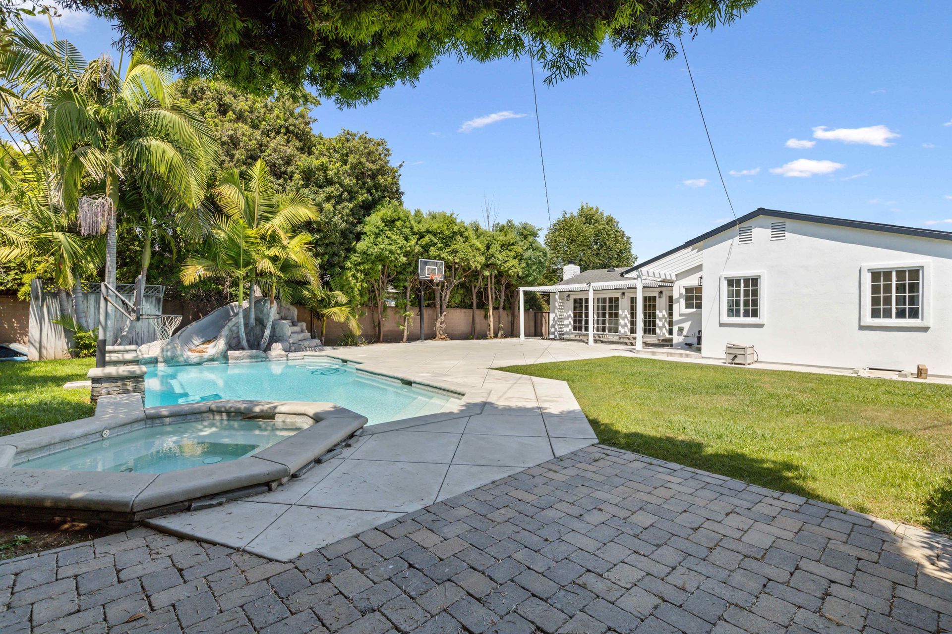 Backyard patio with swimming pool, lawn, and white house under a clear blue sky
