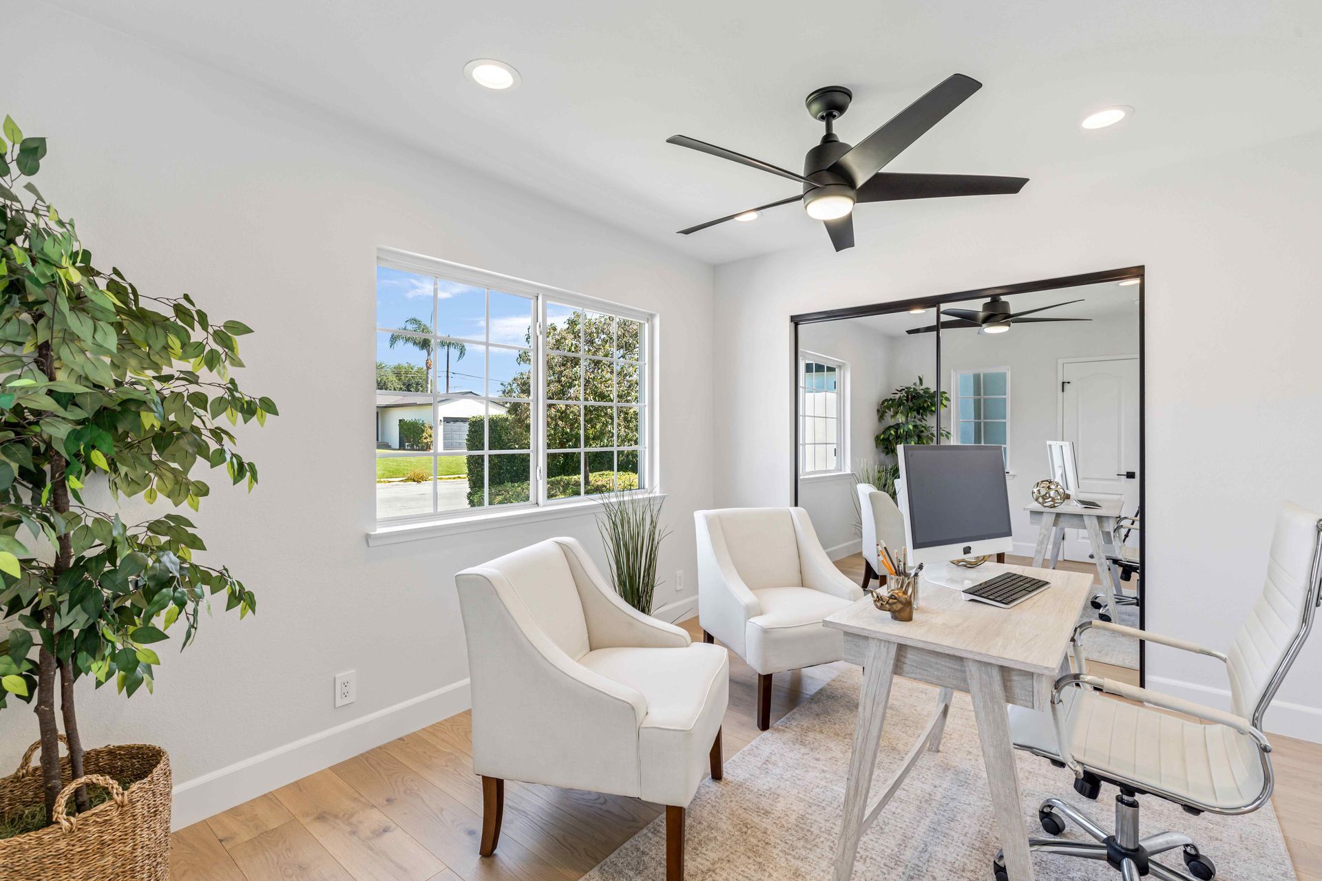 Bright home office with white chairs, desk, large mirror, and ceiling fan by a sunlit window.