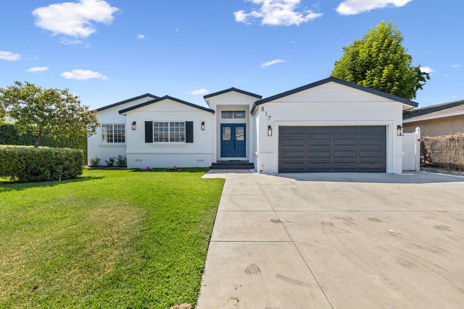 Single-story white house with blue trim, front lawn, and a wide concrete driveway under a clear sky