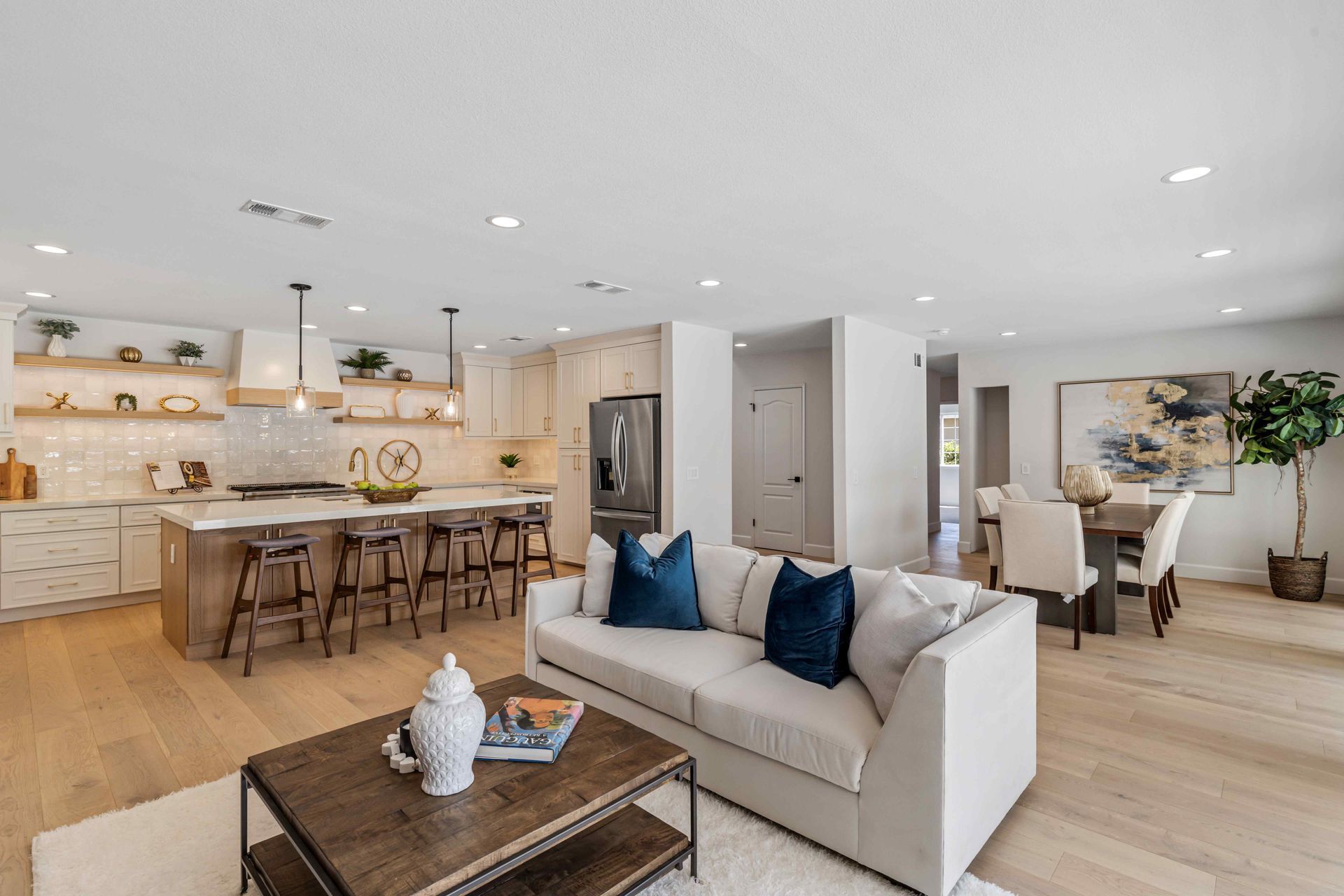 Bright open-plan living room and kitchen with beige sofa, wood floors, bar stools, and white cabinetry