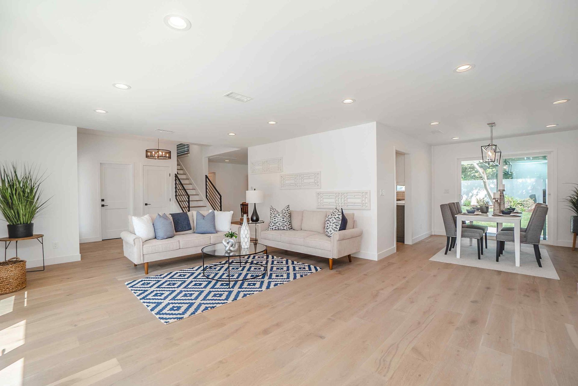 Open-concept living space with two couches, dining table, and staircase. Light wood floors and white walls.