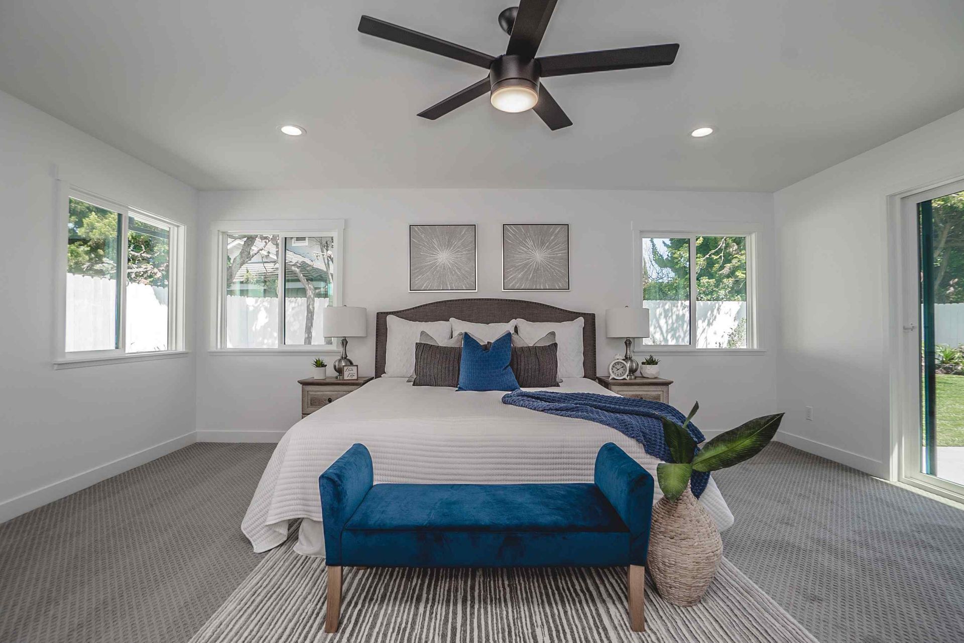 Bedroom with white walls, bed, blue accents, gray carpet, and ceiling fan.
