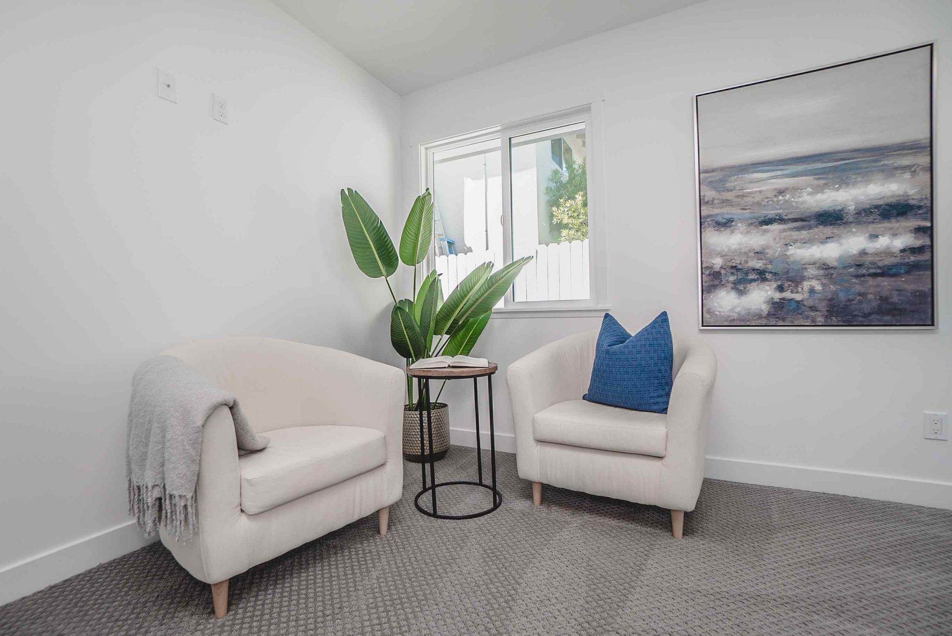 Two cream armchairs face each other in a white-walled room with a plant, small table, and a painting.
