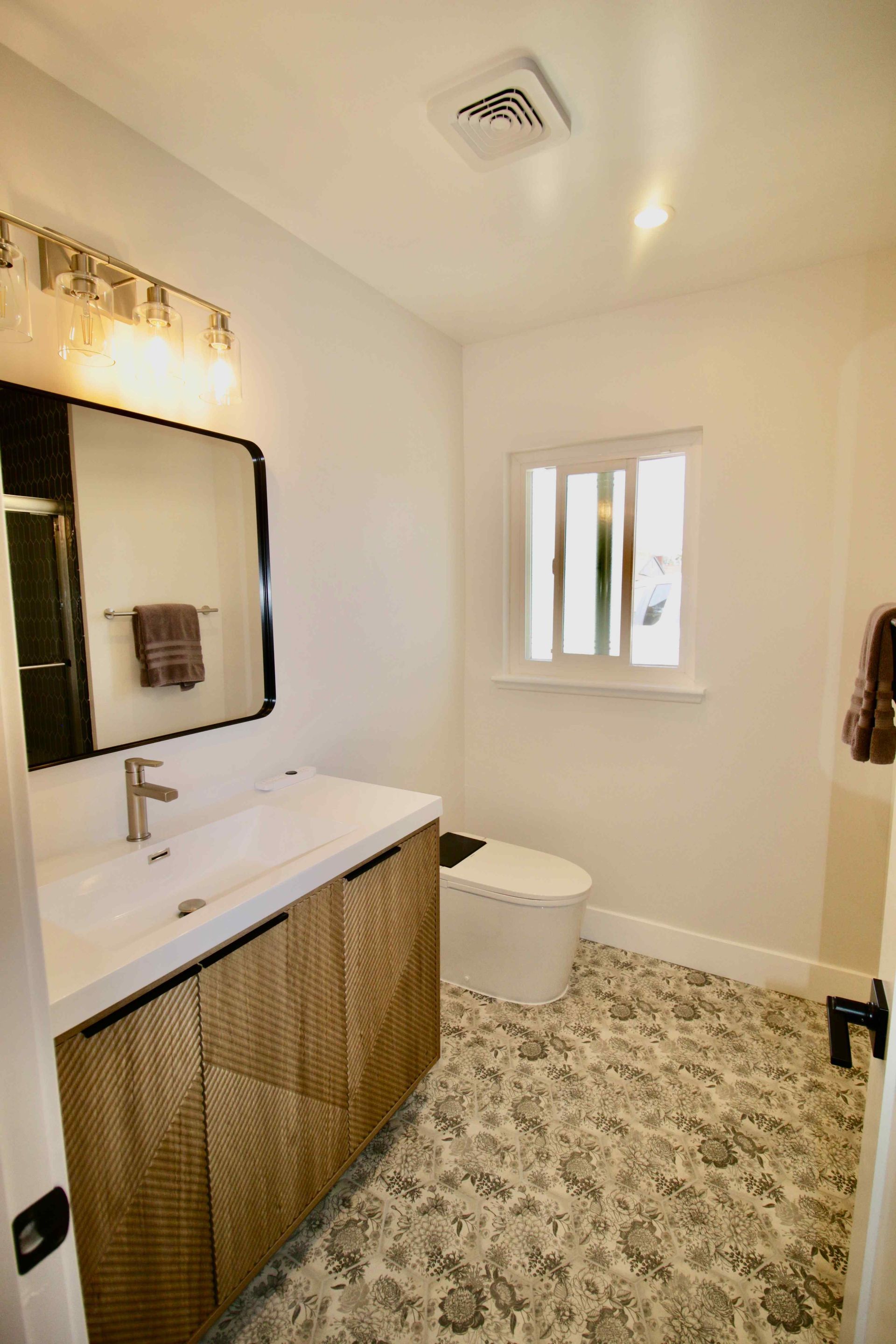 Modern bathroom with floating vanity, large mirror, patterned floor, and small window near the toilet.
