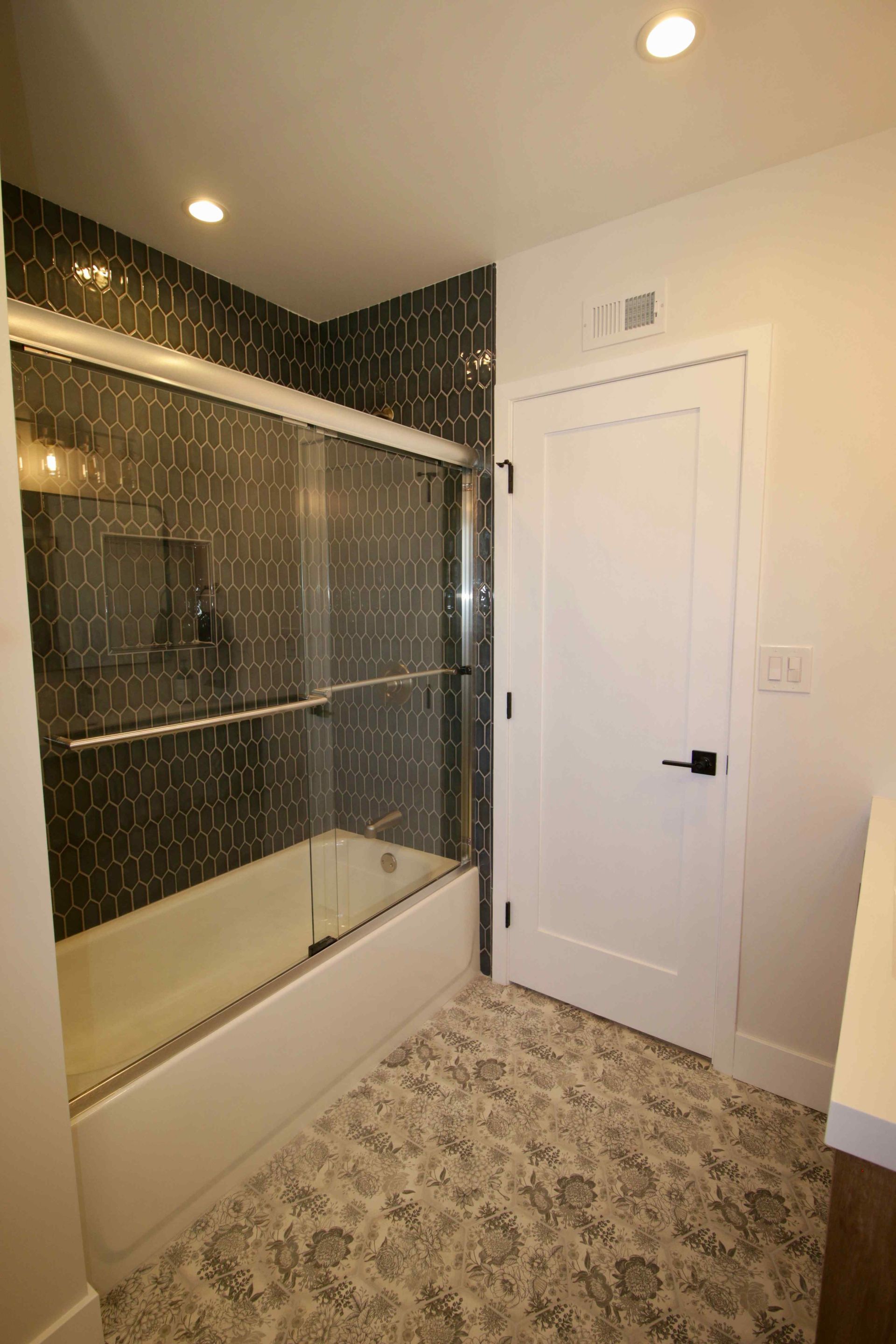 Bathroom with a white door, patterned carpet, and a shower-tub with dark tile surround.