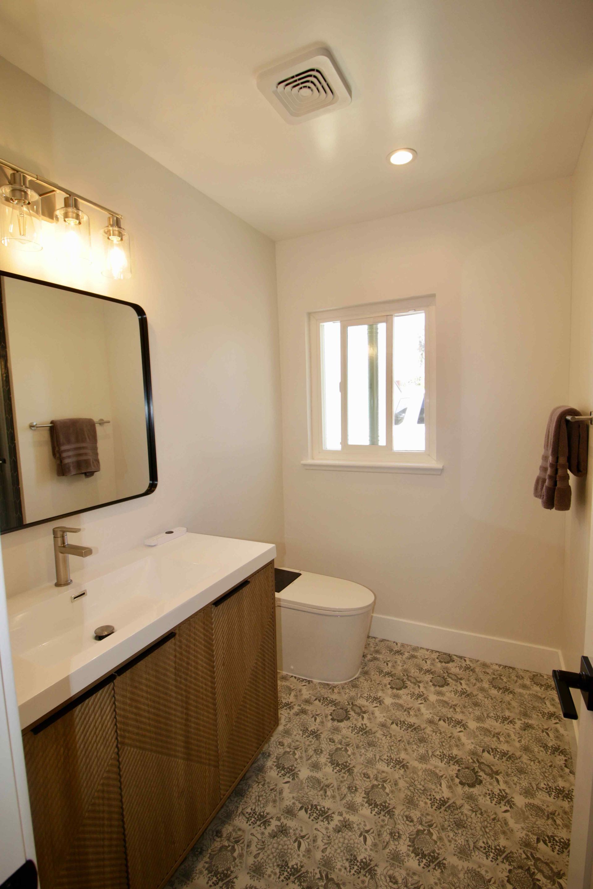Small bathroom with white vanity, mirror, toilet, window, and patterned tile floor.
