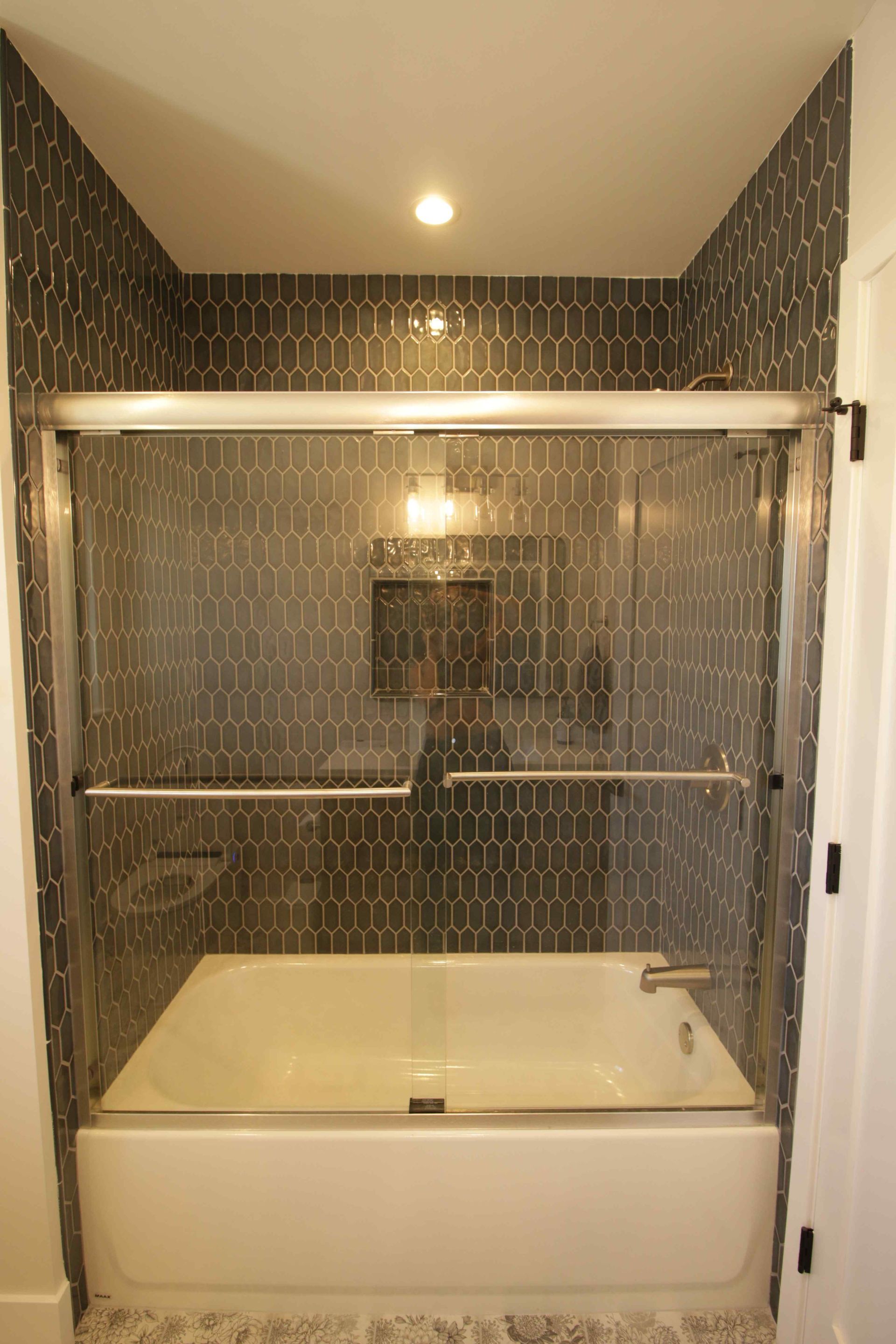 Modern shower alcove with dark patterned tile, glass doors, and white tub base