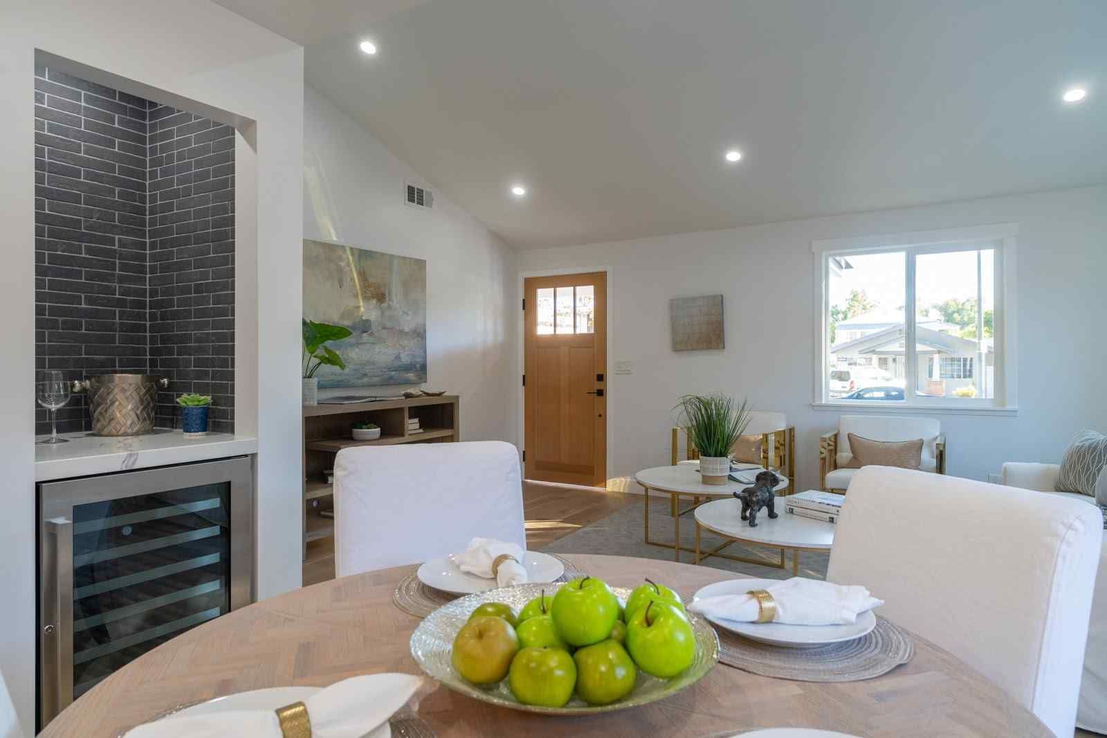 Bright dining area with white chairs, wood table, and bowl of green apples beside a tiled wine cooler.