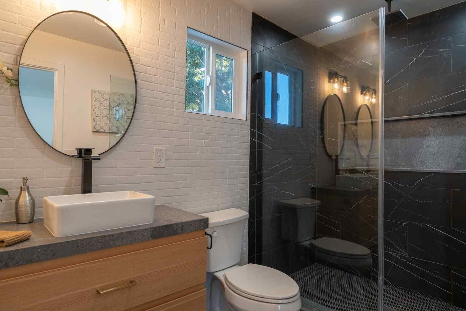 Modern bathroom with wood vanity, round mirror, white sink, toilet, and glass-enclosed shower
