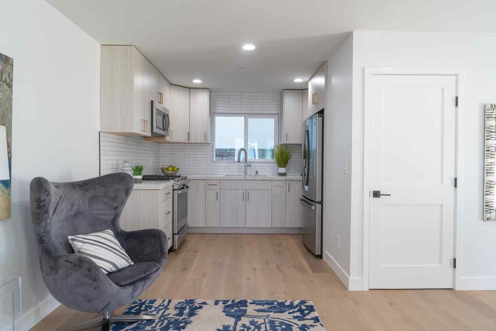 Bright living room opening to a white kitchen with gray chair, blue rug, and light wood floors