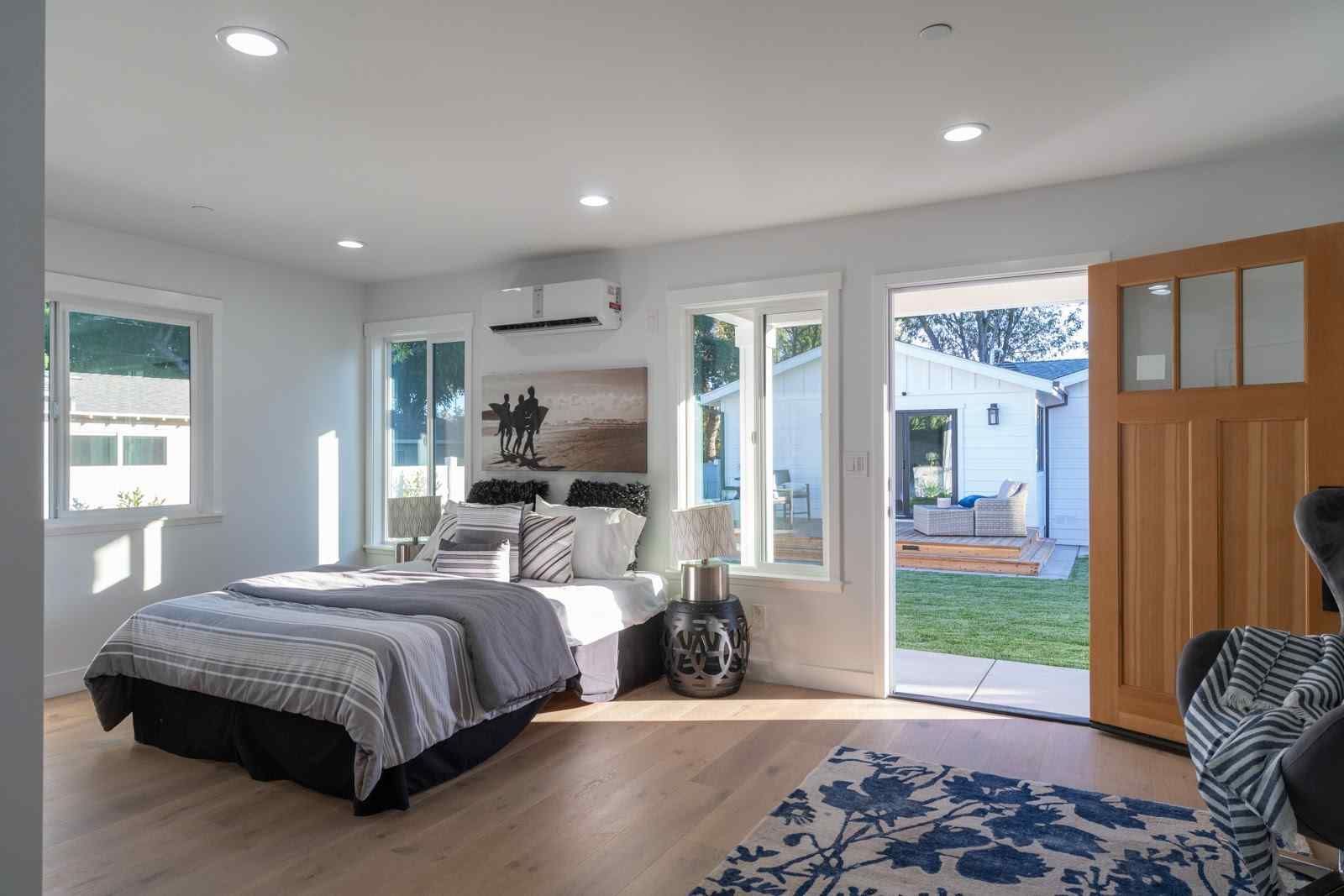 Bright modern bedroom with a bed, windows, open door to the yard, and wood floors.