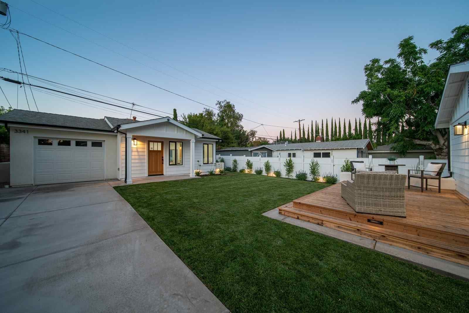 Suburban backyard at dusk with lawn, wooden deck, driveway, garage, and patio seating