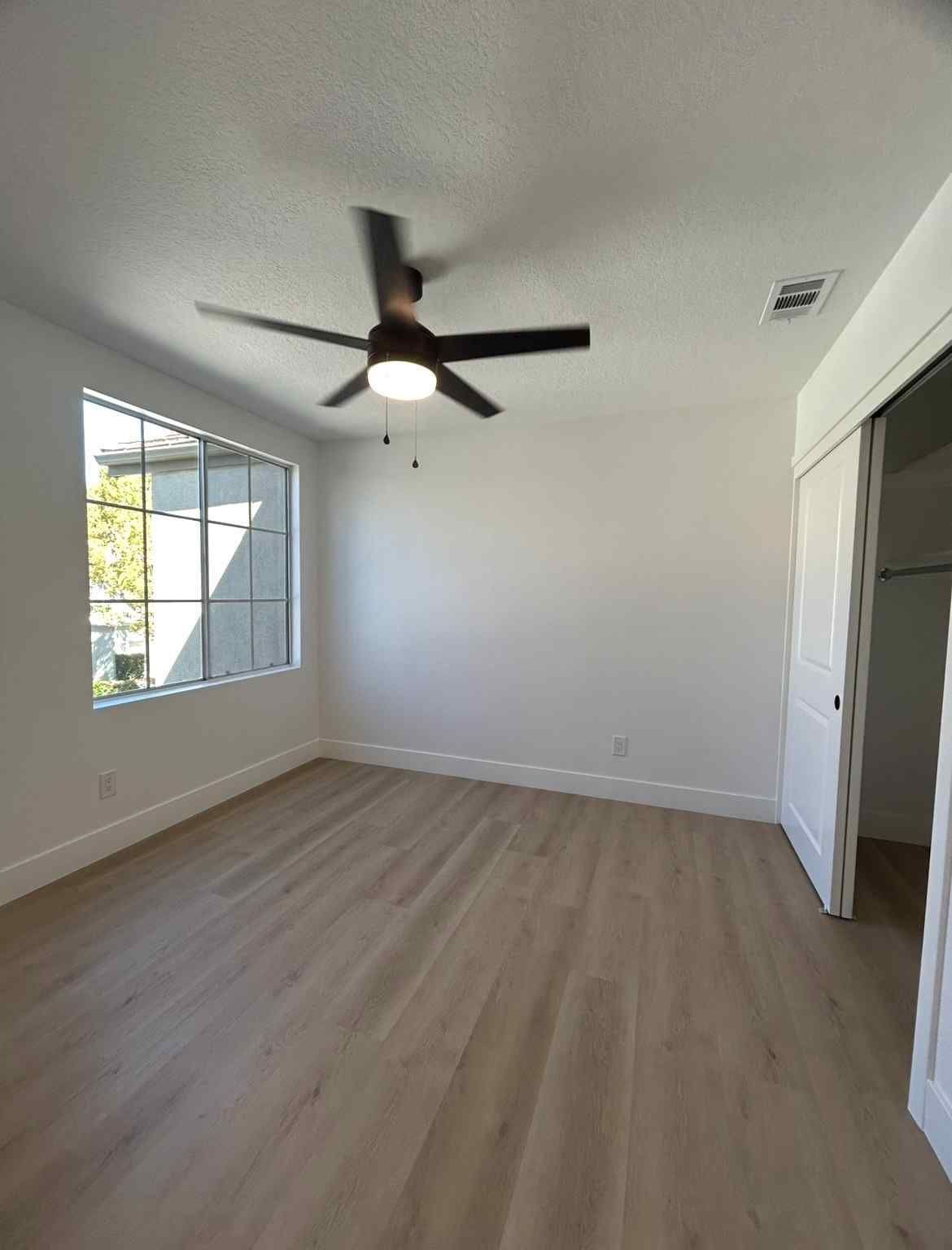 Empty modern bedroom with light wood floor, white walls, ceiling fan, window, and closet door.