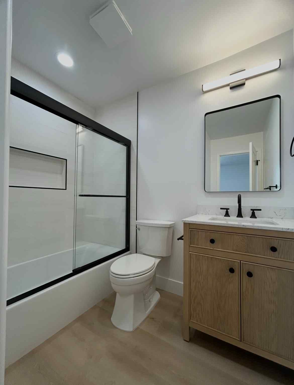 Modern white bathroom with black-framed tub, toilet, and wood vanity with mirror.