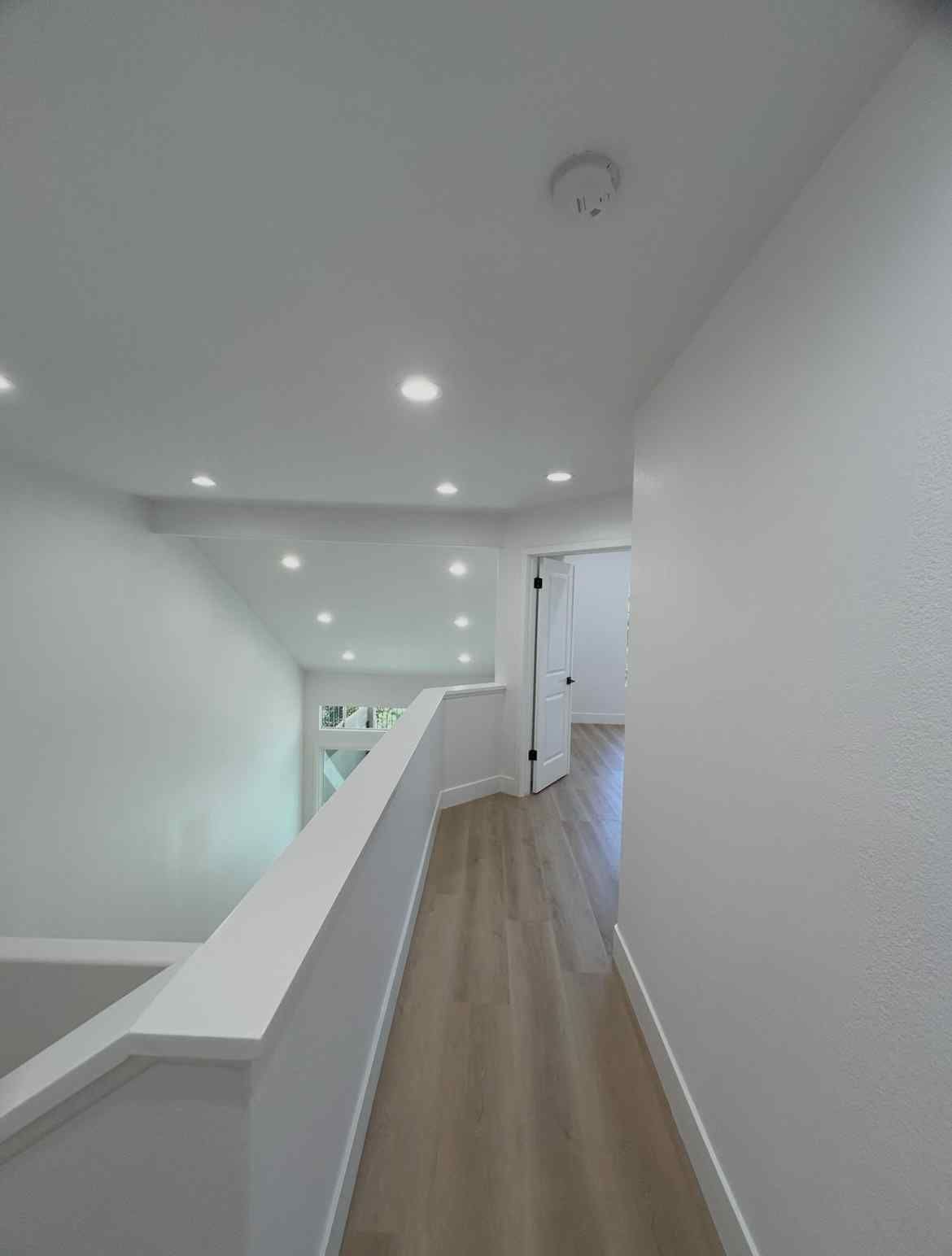 Bright upstairs hallway with white walls, wood floor, and recessed ceiling lights over a stair landing
