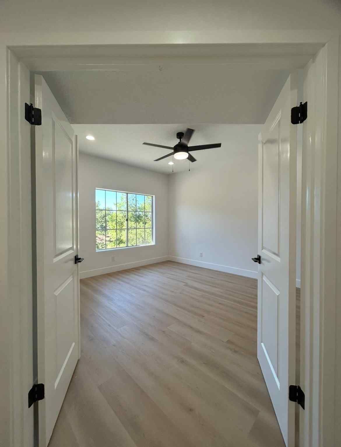 Bright empty bedroom with white walls, wood floors, a ceiling fan, and a window with green trees outside